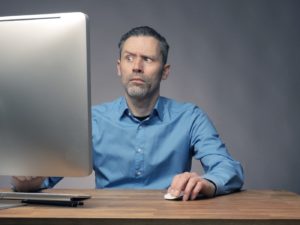 A confused-looking man staring at a computer screen with one eyebrow raised as if he cannot believe what he is seeing.